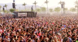 Une image de la foule lors d'un festival de musique, de dos, face à une scène et des palmiers. L'image est floue et prise à distance.
